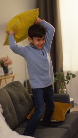 Vertical-Video-Of-Disruptive-Young-Boy-Behaving-Badly-At-Home-Jumping-On-Sofa-And-Throwing-Cushions-Around-Lounge-1